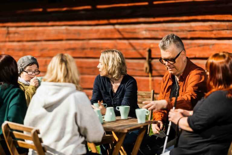 Kuvassa on puutarhapöydän ääressä keskustelemassa ryhmä eri ikäisiä ihmisiä.