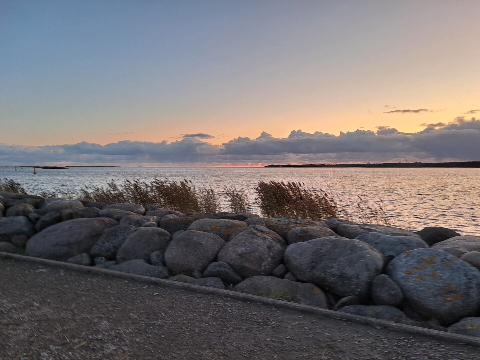 Auringonlaskun sävyinen maisemakuva, jossa kävelytietä reunustaa meren puolella kiviaita.